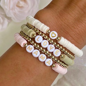 May include: A set of five bracelets with gold beads and white letter beads spelling out "MAMA" and "LOVE". One bracelet has a small gold heart charm.