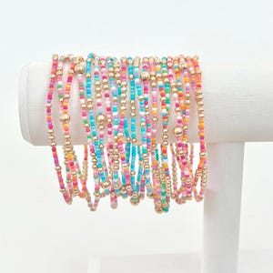 May include: A stack of colorful beaded bracelets with gold beads. The bracelets are arranged in a rainbow pattern and are displayed on a white stand.