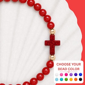 May include: A beaded bracelet featuring smooth, round, glossy red beads and a central red cross charm with a glittery texture. The cross is flanked by two small, polished gold-tone metallic beads. A white rectangular graphic displays the text CHOOSE YOUR BEAD COLOR above a grid of sixteen small, colorful circular bead options.