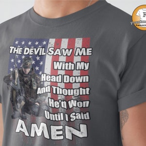 May include: A dark gray t-shirt with a distressed American flag design and the text "THE DEVIL SAW ME With My Head Down And Thought He'd Won Until I Said AMEN".