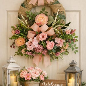 May include: A decorative wreath featuring peach-colored fabric bows, pink peonies, and green foliage, displayed above a wooden sign with the word Welcome, a potted arrangement of pink flowers, a white bird figurine, and two lanterns with candles.