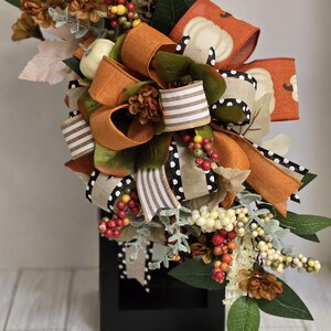 May include: A fall-themed floral arrangement featuring a black base. The arrangement includes orange, green, and white ribbons, faux pumpkins, berries, and various fall foliage. The color palette is dominated by warm autumnal hues.