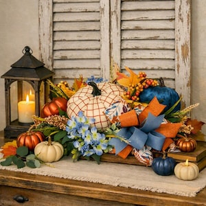 May include: A decorative centerpiece featuring a central white pumpkin with a brown grid pattern, surrounded by smaller metallic copper, cream, and navy blue pumpkins, blue hydrangea flowers, orange and blue ribbons, and a black metal lantern.