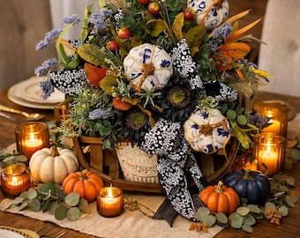 May include: A decorative centerpiece in a wooden basket featuring fabric pumpkins with blue and white patterns, orange pumpkins, dark blue pumpkins, black and white patterned ribbon, faux greenery, dried flowers, and small red berries.