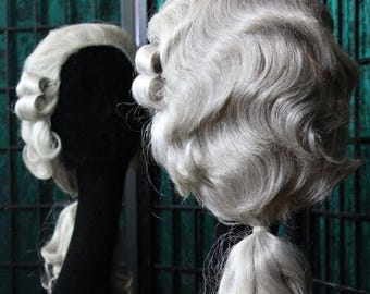 Könnte beinhalten: Zwei graue Perücken mit aufwendigen Locken und einem Pferdeschwanz werden auf schwarzen Schaufensterpuppenköpfen ausgestellt. Die Perücken sind in historischer Mode gestylt, wobei die Locken das Gesicht umrahmen und den Rücken hinunterfallen.