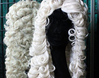 Könnte beinhalten: Zwei weiße, lockige Perücken werden auf schwarzen Schaufensterpuppenköpfen präsentiert. Die Perücken haben ein voluminöses, geschichtetes Design mit engen Locken. Diese Perücken sind wahrscheinlich für Kostüm- oder Theaterzwecke bestimmt.