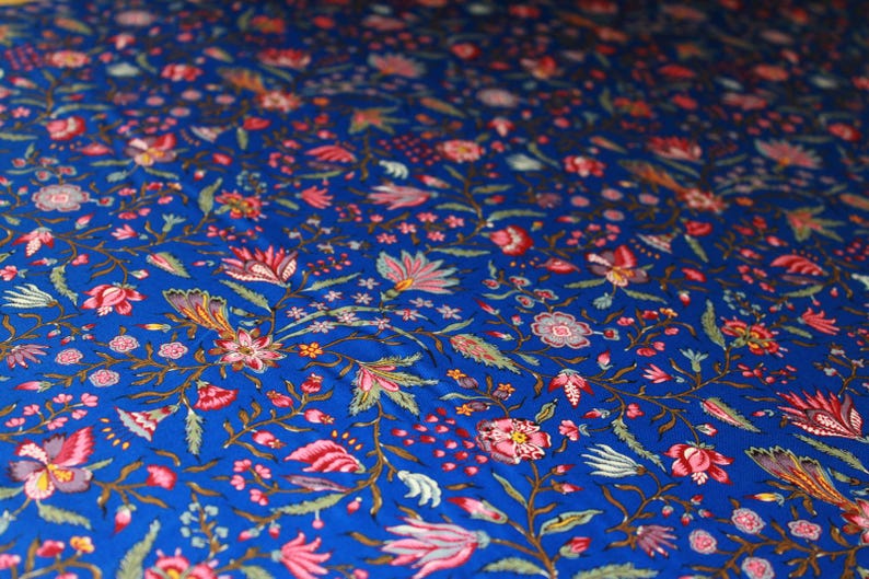 May include: Close-up of a blue fabric with a floral pattern. The design features pink, red, and white flowers with green leaves and stems. The background is a deep blue color, creating a vibrant and detailed textile.