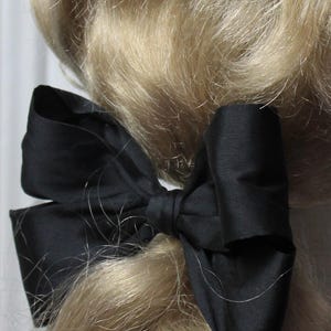 Può includere: Un accessorio per capelli a fiocco in raso nero. Il fiocco è legato e attaccato ai capelli biondi. Il fiocco è di colore nero pieno ed è realizzato in un materiale lucido e liscio. I capelli sono biondo chiaro e ondulati.