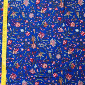 May include: Vibrant blue fabric with a detailed floral pattern in shades of pink, red, and green. The design features stylized flowers, leaves, and small star-shaped accents. A yellow measuring tape is visible along the left edge, showing measurements in inches.