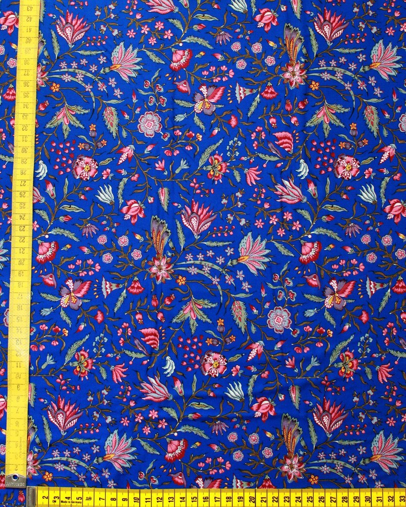 May include: Vibrant blue fabric with a detailed floral pattern in shades of pink, red, and green. The design features stylized flowers, leaves, and small star-shaped accents. A yellow measuring tape is visible along the left edge, showing measurements in inches.