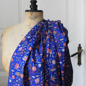 May include: A bolt of vibrant blue fabric draped over a mannequin. The fabric is densely patterned with a floral design in shades of red, orange, and white. The mannequin is a light beige color, and the background is a white door.