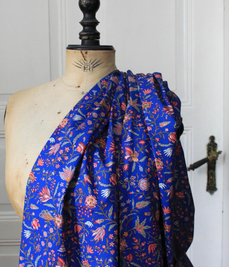 May include: A bolt of vibrant blue fabric draped over a mannequin. The fabric is densely patterned with a floral design in shades of red, orange, and white. The mannequin is a light beige color, and the background is a white door.