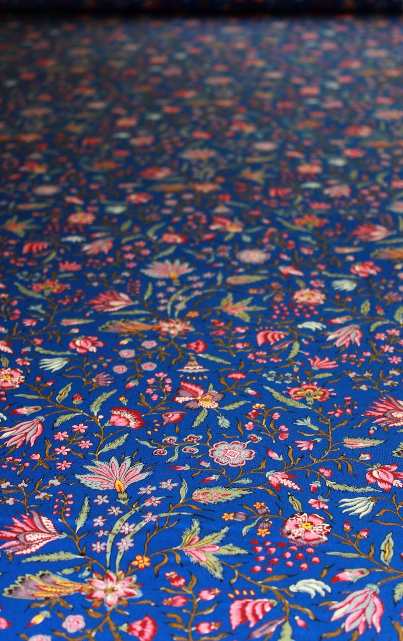 May include: Close-up of a vibrant blue fabric with a repeating floral pattern. The design features pink, red, and green flowers with delicate leaves and stems. The fabric is likely used for textiles or home decor.