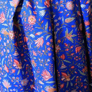 May include: Close-up of a vibrant blue fabric with a detailed floral pattern. The design features red, pink, and white flowers with green leaves and stems. The fabric is folded, creating shadows and depth, highlighting the intricate print.