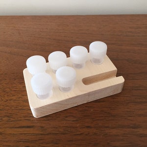 Puede incluir: Una paleta de pintura de madera con seis pequeños recipientes de plástico transparente con tapas blancas. La paleta es de madera clara y tiene una ranura en un lado. Los recipientes están dispuestos en dos filas de tres.