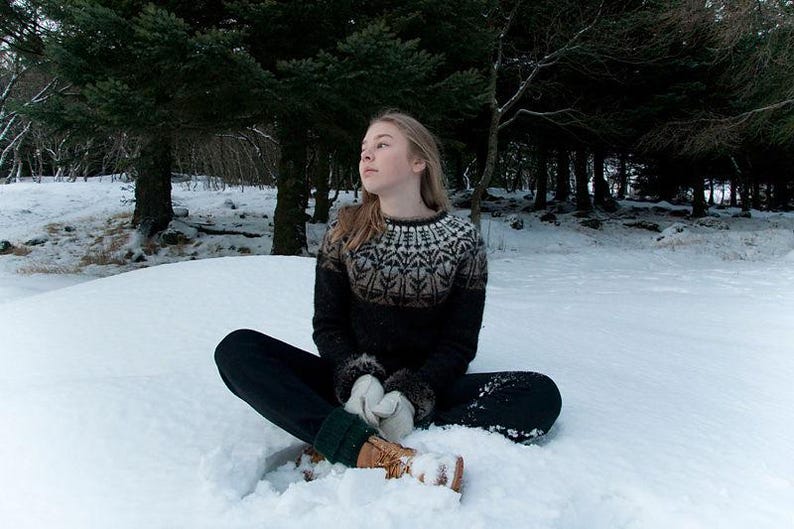 May include: A person wearing a black and white patterned sweater, black pants, and brown boots sits cross-legged in the snow. The person is wearing white gloves and is surrounded by snow and trees.
