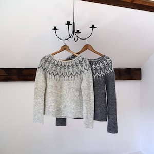 May include: Two sweaters hang from wooden hangers. The sweaters are gray and white with patterned designs around the neckline. A black chandelier hangs above the sweaters. The sweaters are displayed against a white wall.