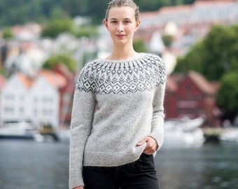 Könnte beinhalten: Ein hellgrauer Pullover mit einem geometrischen Muster am Halsausschnitt. Der Pullover hat lange Ärmel und wird mit einer schwarzen Jeans kombiniert. Der Hintergrund zeigt ein Gewässer und Gebäude.
