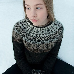 May include: A person wearing a black and brown patterned sweater with a round neckline. The sweater has a geometric design around the yoke. The person is wearing black pants and white gloves with fur trim. The background is a snowy landscape.