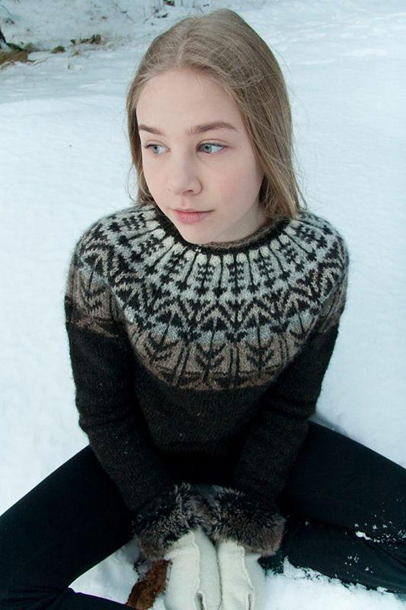 May include: A person wearing a black and brown patterned sweater with a round neckline. The sweater has a geometric design around the yoke. The person is wearing black pants and white gloves with fur trim. The background is a snowy landscape.