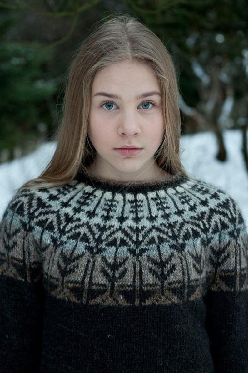 May include: A person wearing a patterned sweater with a black base and a detailed design in shades of gray, brown, and white. The sweater has a round neckline and long sleeves. The background is a blurred winter scene.