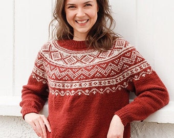 May include: A rust-colored sweater with a cream-colored geometric pattern across the chest and shoulders. The sweater has a crew neck and long sleeves. The person in the image is smiling and leaning against a white wall.