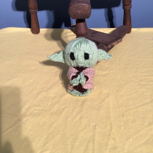 Puede incluir: Un juguete de peluche verde tejido de un personaje popular de la franquicia Star Wars. El juguete lleva una chaqueta de punto rosa y tiene un cuello de punto marr&oacute;n.