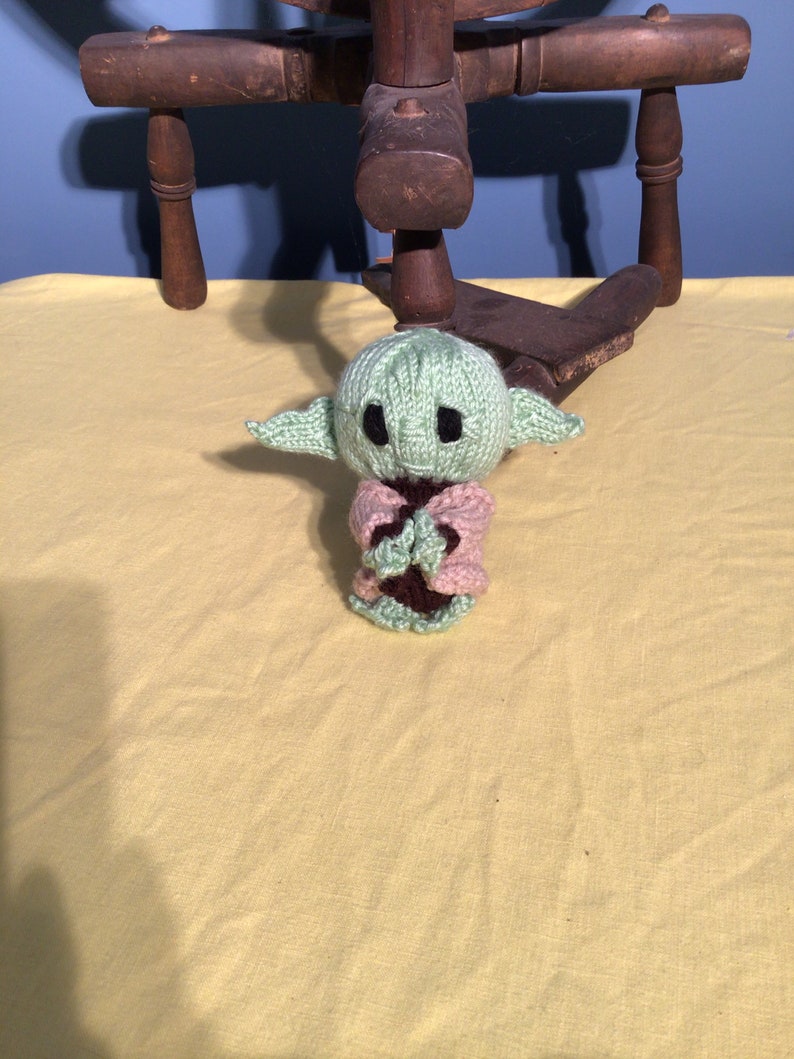 Puede incluir: Un juguete de peluche verde tejido de un personaje popular de la franquicia Star Wars. El juguete lleva una chaqueta de punto rosa y tiene un cuello de punto marr&oacute;n.