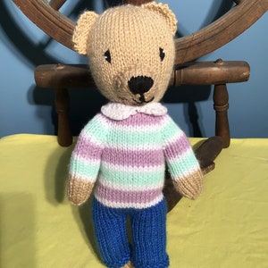 Puede incluir: Un oso de peluche tejido que lleva un su&eacute;ter a rayas y pantalones vaqueros azules. El oso tiene una nariz marr&oacute;n y ojos negros.