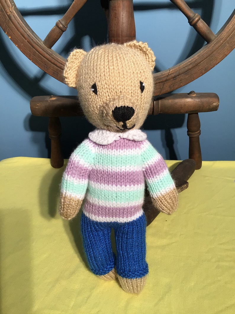 Puede incluir: Un oso de peluche tejido que lleva un su&eacute;ter a rayas y pantalones vaqueros azules. El oso tiene una nariz marr&oacute;n y ojos negros.