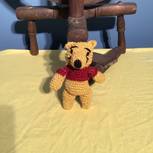 Puede incluir: Un oso de peluche Winnie the Pooh de crochet con una camisa roja. El oso est&aacute; de pie sobre un mantel amarillo.