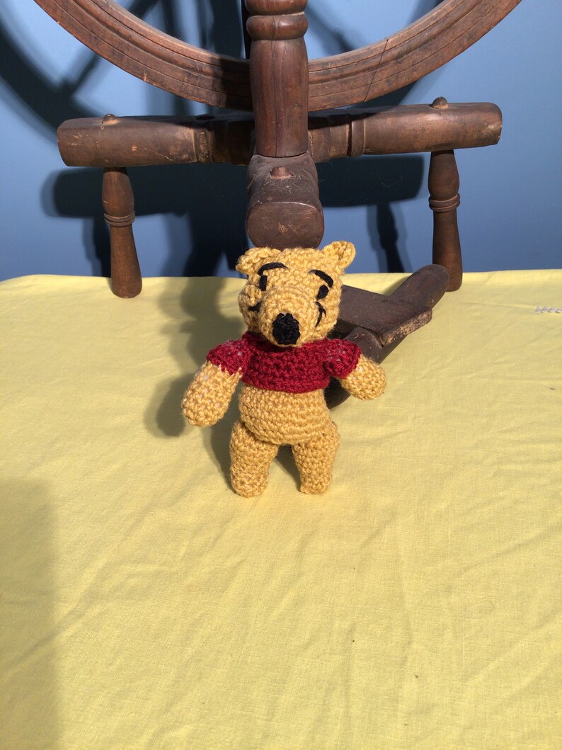 Puede incluir: Un oso de peluche Winnie the Pooh de crochet con una camisa roja. El oso est&aacute; de pie sobre un mantel amarillo.