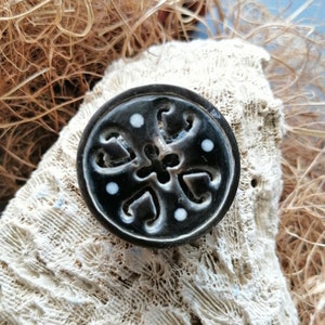 Könnte beinhalten: Ein schwarzer Keramik-Knopf mit einem weißen Blumenmuster und weißen Punkten. Der Knopf liegt auf einer weißen korallenartigen Oberfläche.