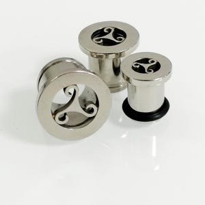 Op de afbeelding: Drie zilveren metalen oorpluggen met een Keltisch knoopontwerp. De pluggen zijn in verschillende maten.