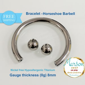 Op de afbeelding: Een zilveren hoefijzer barbell armband gemaakt van hypoallergeen titanium. De armband is 8 mm dik en heeft twee zilveren ballen aan de uiteinden. De armband is gelabeld "Nickel free Hypoallergenic Titanium" en "Gauge thickness (0g) 8mm".