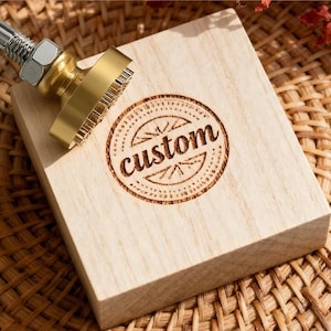 May include: A wooden stamp with the word "custom" engraved in a circular design. The stamp has a brass head and a metal handle. The wooden block is light-colored and sits on a woven surface.