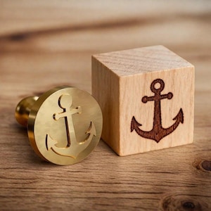 May include: A brass anchor stamp with a wooden handle and a wooden block with an anchor design. The stamp is round with a detailed anchor cutout. The wooden block has a dark brown anchor image.