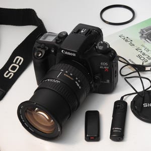 May include: A black Canon EOS Elan 7E camera with a zoom lens, strap, and accessories. The camera has a lens attached, a lens cap, a remote control, and a manual. The strap is black with the Canon and EOS logos in white.