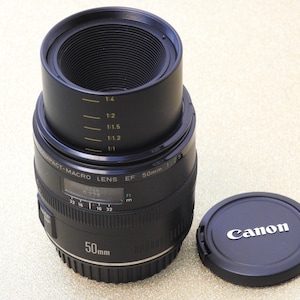 May include: A black Canon 50mm macro lens with a lens hood and lens cap. The lens has markings for focus distance and aperture settings. The lens cap is black with the Canon logo in white.