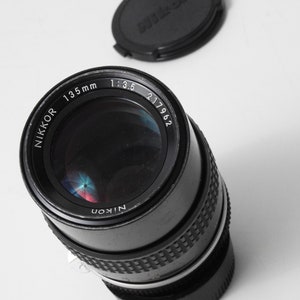 May include: A black Nikon Nikkor 135mm f/3.5 camera lens with a matching lens cap. The lens has a silver ring and the text "NIKKOR 135mm 1:3.5 217962" is visible. The lens cap is black and has the Nikon logo.