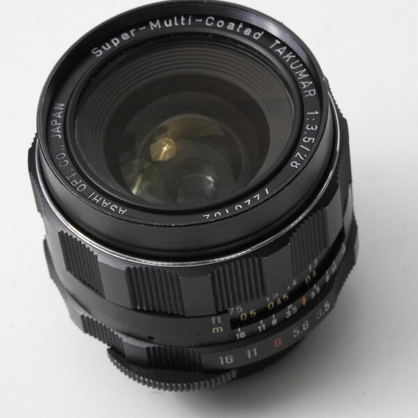 以下が含まれることがあります： 黒色のSuper-Multi-Coated Takumar 1:3.5/28カメラレンズ。レンズには「ASAHI OPT. CO. JAPAN」や「7010771」などの数字やマーキングが施されたリングがあります。写真撮影用に設計されています。