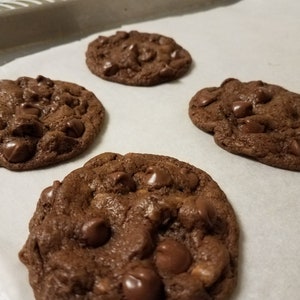 May include: Four chocolate chip cookies on a baking sheet lined with parchment paper. The cookies are slightly browned and have a soft, chewy texture.