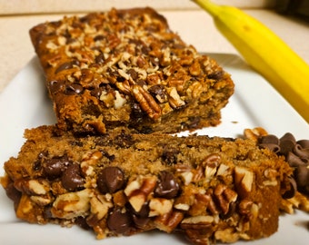 Può includere: Una pagnotta di banana bread con gocce di cioccolato e noci pecan su un piatto bianco. Il pane è tagliato a fette e la parte superiore è ricoperta di noci pecan.