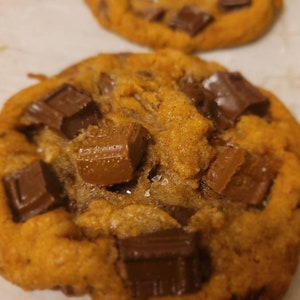 May include: A close-up of a chocolate chip cookie with large pieces of milk chocolate on top. The cookie is golden brown and has a slightly crispy texture.