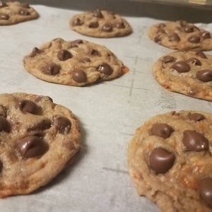 May include: A baking sheet lined with parchment paper and filled with freshly baked chocolate chip cookies. The cookies are golden brown and have a variety of sizes and shapes.