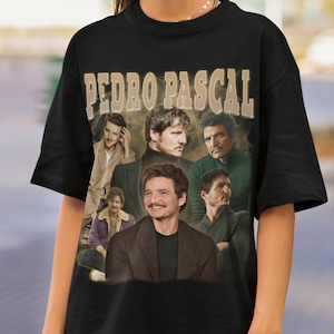 Könnte beinhalten: Schwarzes T-Shirt mit einem Vintage-Grafikdruck, der eine Collage von Bildern des Schauspielers Pedro Pascal zeigt. Der Text "PEDRO PASCAL" ist in goldenen Buchstaben über der Collage gedruckt.