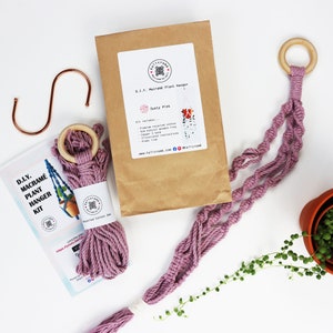 Könnte beinhalten: Ein staubiges rosa Makramee-Pflanzenhänger-Set mit einem Holzring, Baumwollschnur und Anleitungen. Das Set enthält einen Kupferhaken und einen Videolink. Das Set ist in einer braunen Papiertüte verpackt.
