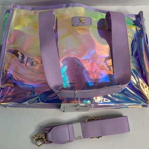May include: A rectangular tote bag with an iridescent, holographic finish and lavender trim. It features two lavender fabric handles, a matching detachable shoulder strap with gold-tone metal clips, and a small rectangular patch with text.