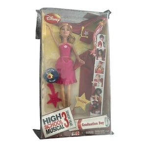 Puede incluir: Muñeca Disney High School Musical en un embalaje de plástico transparente. La muñeca lleva un vestido rosa y tiene el pelo rubio. Los accesorios incluyen un charm en forma de estrella, un pergamino de graduación y un pin "Class ring for you!".