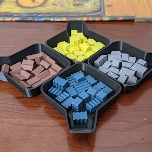 May include: Four black plastic trays with raised edges, each containing a different color of small plastic building blocks. The trays are arranged in a square shape, with the yellow blocks in the top tray, the blue blocks in the bottom tray, the brown blocks in the left tray, and the gray blocks in the right tray.